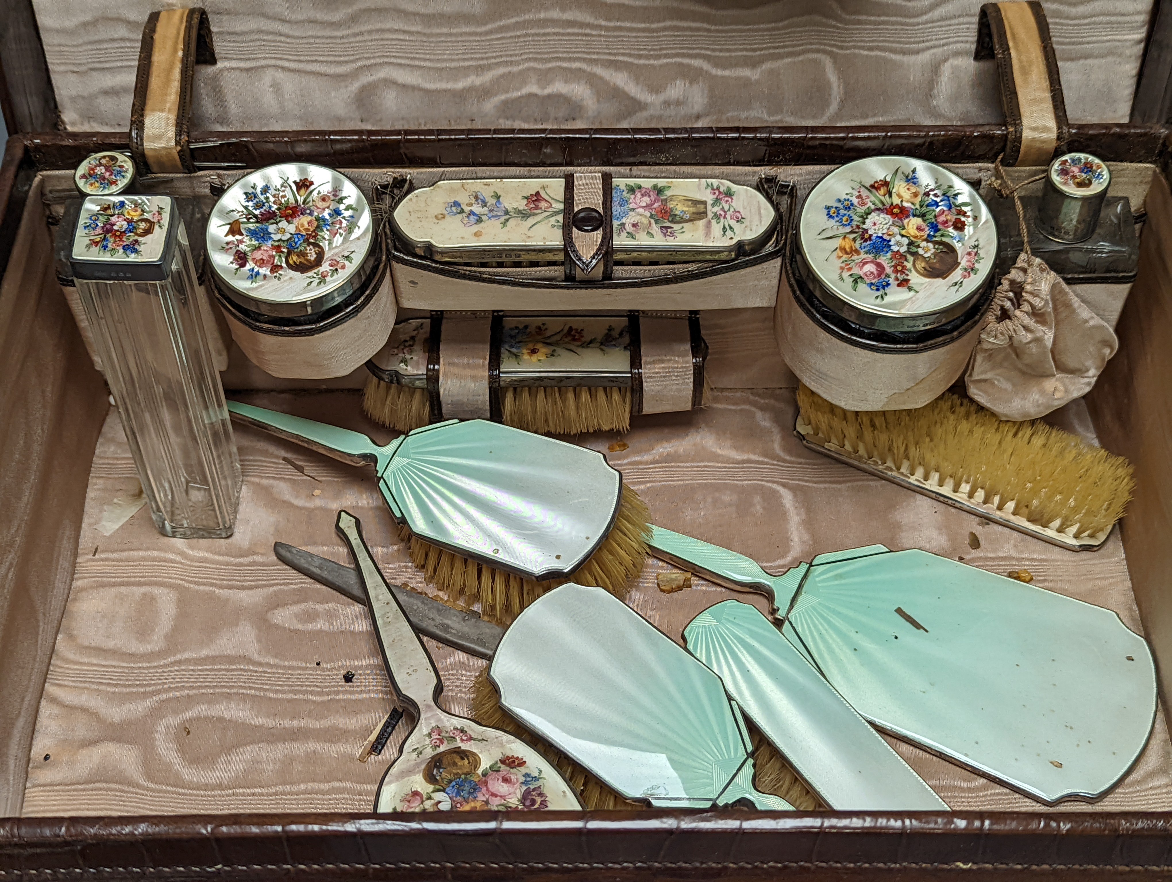 A 1930's crocodile skin travelling toilet case, with ten silver and floral enamel mounted accoutrements, Henry Clifford Davis, Birmingham, 1935,6, together with five other silver and enamel brushes and mirror etc and a c